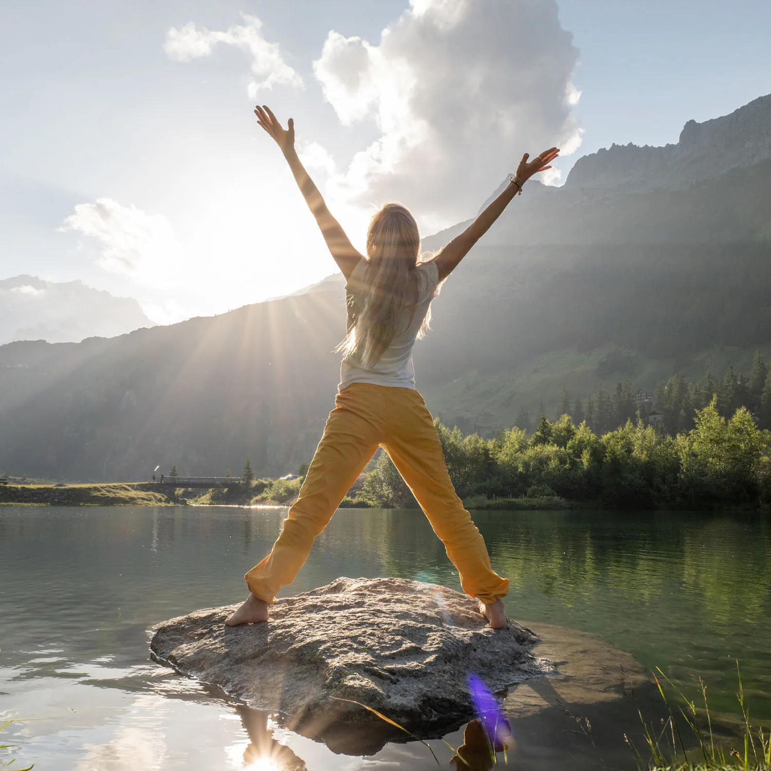 Eine Frau steht mit ausgestreckten Armen auf einem Stein im See und wird von der Sonne angestrahlt.